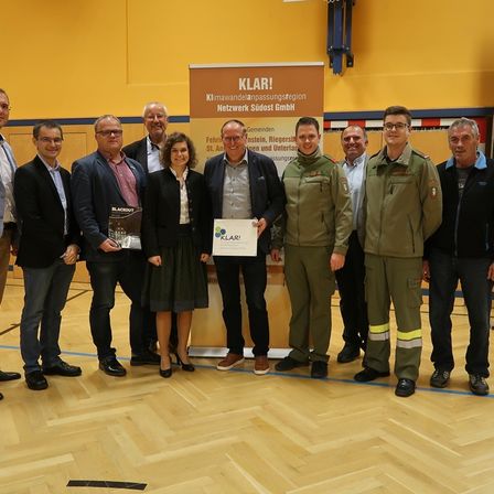 Eine Gruppe von Menschen steht nebeneinander und hält Urkunden in einem Raum mit einer Projektionswand.