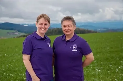 Zwei Frauen stehen in einem Feld mit Bergen im Hintergrund. Sie tragen lila Poloshirts.