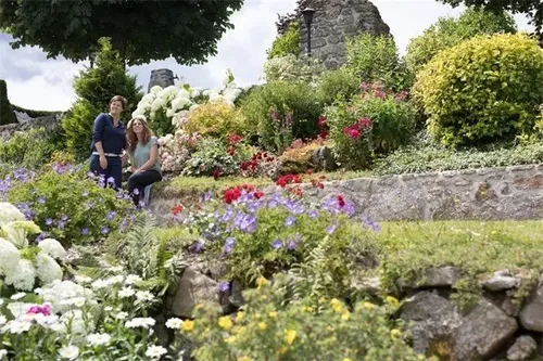 Zwei Frauen posieren für ein Foto in einem Garten voller verschiedener Blumen, Büschen und Bäumen, mit Steinmauern und einer Ziegelstruktur im Hintergrund.