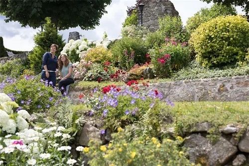 Zwei Frauen posieren für ein Foto in einem Garten voller verschiedener Blumen, Büschen und Bäumen, mit Steinmauern und einer Ziegelstruktur im Hintergrund.