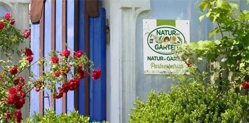 Ein weißes Schild mit blauen und grünen Akzenten ist an einer blau-weißen Wand befestigt. Das Schild trägt die Aufschrift 'NATUR-GARTEN Natur-Garten Partnerschaft'. Im Vordergrund befinden sich rote Blumen und grüne Büsche.
