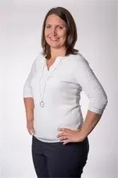 Eine Frau in einem weißen Langarmshirt und schwarzen Hosen lächelt und hat die Hände auf den Hüften.