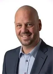 Ein Mann mit kahlem Kopf trägt einen Anzug und ein hellblaues Hemd. Er lächelt und posiert für ein Foto.