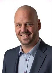 Ein Mann mit kahlem Kopf trägt einen Anzug und ein hellblaues Hemd. Er lächelt und posiert für ein Foto.
