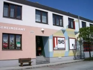 Ein Gemeindeamtsgebäude mit rosa Fassade, weißen und blauen geometrischen Mustern und einem Schild mit der Aufschrift '1945'. Vor dem Gebäude steht eine Bank und ein Baum.