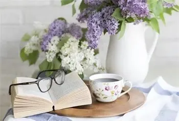 Ein offenes Buch, eine Tasse Tee und eine Brille liegen auf einem runden Holztablett, mit einer weißen Vase mit lila Blumen im Hintergrund.