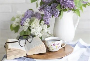 Ein offenes Buch, eine Tasse Tee und eine Brille liegen auf einem runden Holztablett, mit einer weißen Vase mit lila Blumen im Hintergrund.