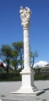 Eine weiße Marmorsäule mit einer religiösen Skulptur einer Familie auf der Spitze, platziert in einem Außenbereich mit einem Ziegelboden, umgeben von einem Zaun und Bäumen im Hintergrund.