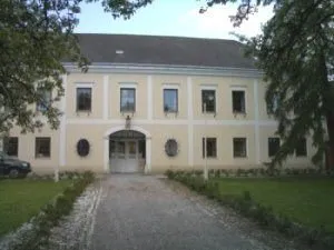 Ein beiges Gebäude mit mehreren Fenstern und einem grauen Dach. Eine Einfahrt führt zum Eingang mit zwei Rundbogenfenstern und einer Lampe über der Tür.