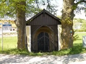 Ein kleines Holzgebäude, möglicherweise eine Kapelle, steht zwischen zwei großen Bäumen mit einem Bogeneingang und einem geschlossenen Tor.