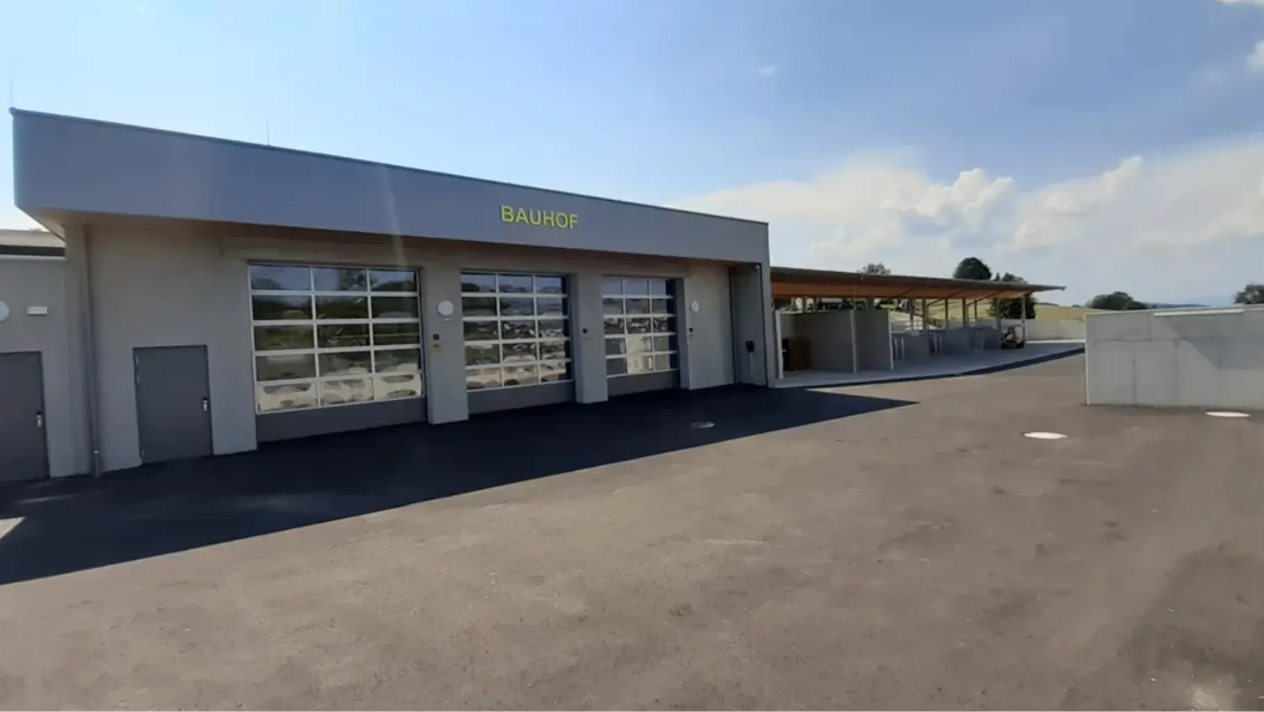 Die Außenansicht eines Gebäudes mit dem Wort Bauhof auf dem Dach. An der Vorderseite des Gebäudes befinden sich große Glastüren und davor ein gepflasterter Parkplatz.
