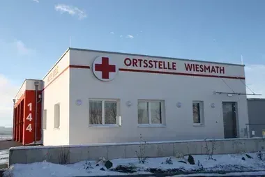 Ein weißes Gebäude mit rotem Kreuzsymbol und der Aufschrift 'Ortsstelle Wiesmath' oben. Es hat drei Fenster und eine Tür. Die Nummer '144' ist an der Wand neben dem Gebäude gemalt. Schnee bedeckt den Boden davor.