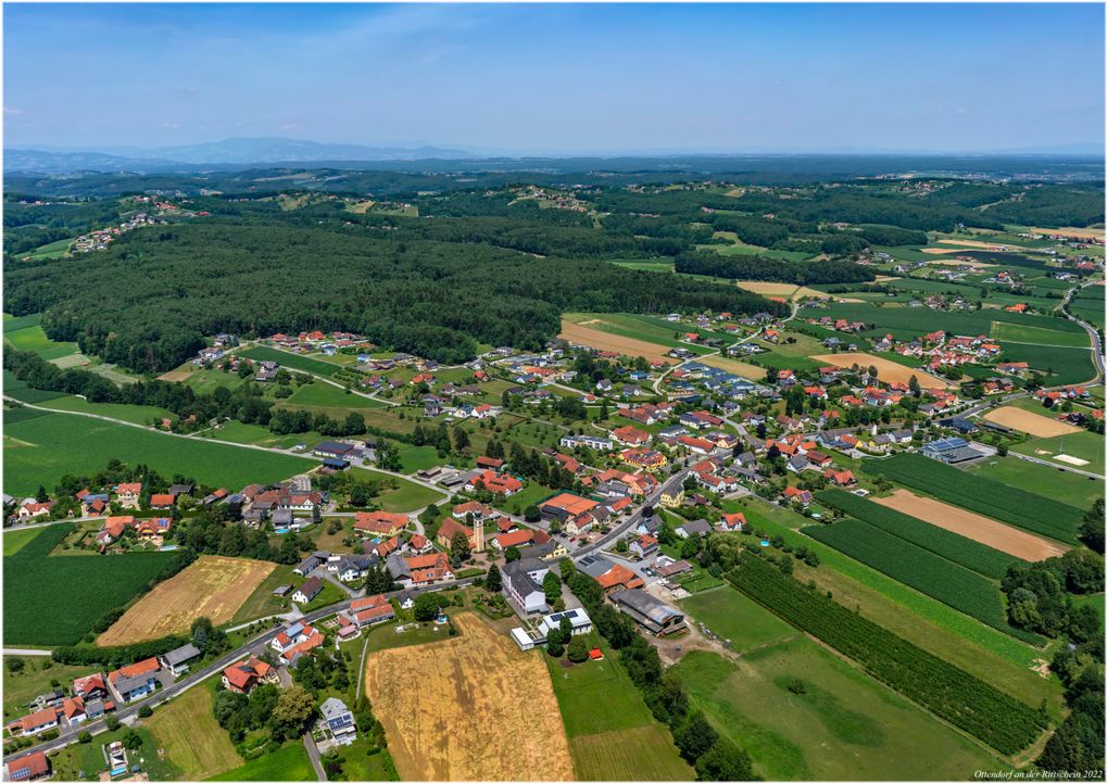  Abschnitt Bild: Luftaufnahme eines Dorfes, umgeben von üppigen grünen Feldern und Bäumen unter einem klaren blauen Himmel. Das Dorf ist mit Häusern in verschiedenen Farben übersät, einige mit roten Dächern. In der Ferne sind Berge sichtbar.