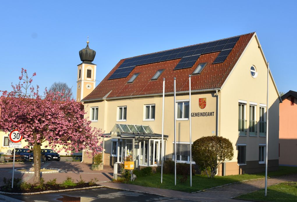  Abschnitt Bild: Ein gelbes Gebäude mit Solarpanelen auf dem Dach, beschriftet mit 'Gemeindeamt', steht neben einem Turm mit einer Uhr und einem rosa blühenden Baum.