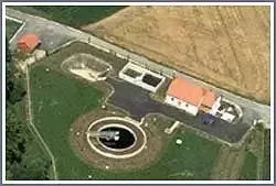Luftaufnahme eines ländlichen Anwesens mit einem großen runden Teich, einem rotgedeckten Haus, einer Einfahrt und einer Scheune auf einem grasbewachsenen Feld.