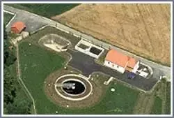 Luftaufnahme eines ländlichen Anwesens mit einem großen runden Teich, einem rotgedeckten Haus, einer Einfahrt und einer Scheune auf einem grasbewachsenen Feld.