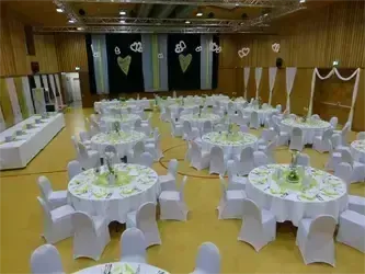Ein großer Innenbereich ist mit vielen runden Tischen und Stühlen eingerichtet. Die Tische sind mit weißen Tischdecken und grünen Blumenarrangements bedeckt. Die Wände sind mit schwarzen und weißen Kulissen und weißen Vorhängen dekoriert.