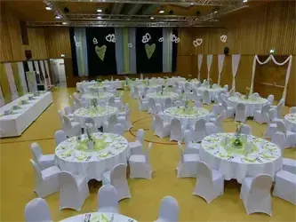 Ein großer Innenbereich ist mit vielen runden Tischen und Stühlen eingerichtet. Die Tische sind mit weißen Tischdecken und grünen Blumenarrangements bedeckt. Die Wände sind mit schwarzen und weißen Kulissen und weißen Vorhängen dekoriert.