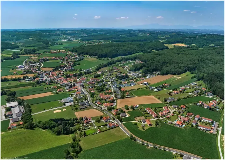 Luftaufnahme eines ländlichen Gebiets mit Häusern, Straßen und Feldern. Grüne Felder umgeben das Gebiet, und eine Straße verläuft durch die Mitte. Der Himmel ist klar mit einigen Wolken.