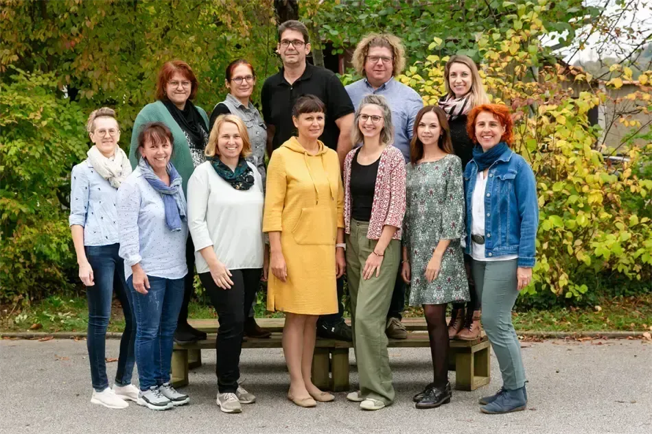 Eine Gruppe von Menschen, bestehend aus Frauen und Männern, steht und sitzt zusammen vor einer Bank. Sie alle lächeln und posieren für ein Foto.