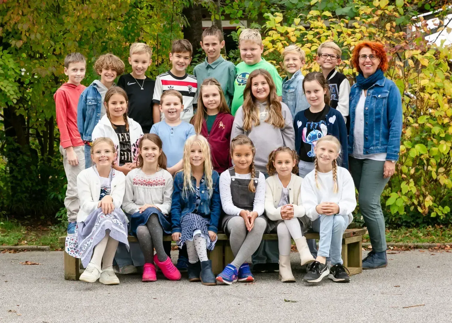 Eine Gruppe junger Kinder, sowohl Jungen als auch Mädchen, posiert für ein Foto im Freien. Sie sitzen auf einer Bank mit Bäumen im Hintergrund.