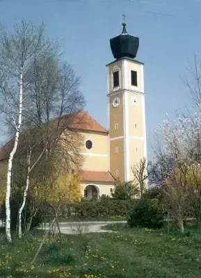 Eine Kirche mit einem hohen Turm und einer Uhr, umgeben von Bäumen und Gras in einem ländlichen Gebiet.