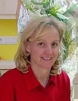 Eine Frau mit blonden Haaren in einem roten Poloshirt lächelt und steht vor einer gelben Wand mit einer Pflanze.