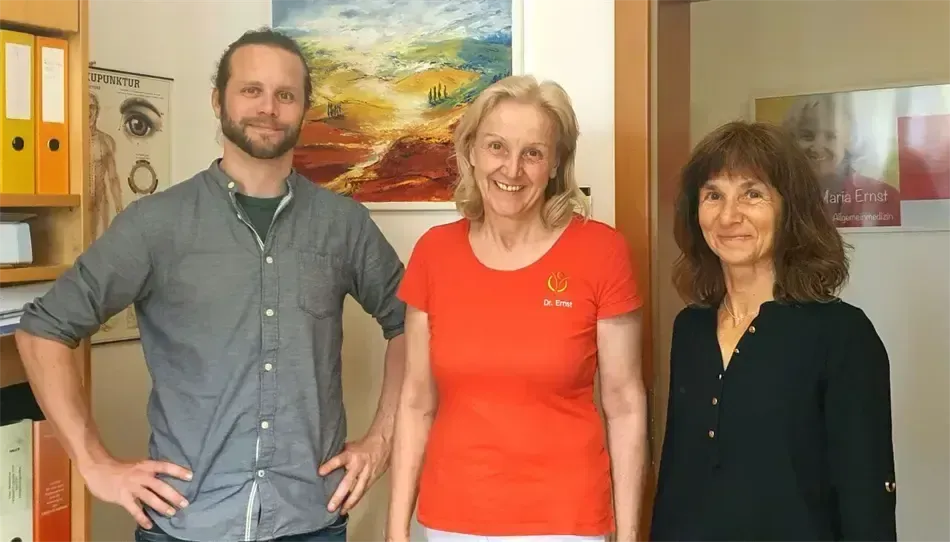 Drei Menschen stehen nebeneinander und lächeln vor einer weißen Wand. Der Mann links hat einen Bart und trägt ein graues Hemd. Die Frau in der Mitte trägt ein orangefarbenes T-Shirt mit Logo. Die Frau rechts trägt ein schwarzes Hemd.