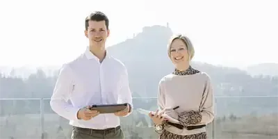 Ein Mann und eine Frau stehen auf einem Balkon mit einem Berg im Hintergrund, lächeln und schauen in die Kamera. Der Mann hält ein Tablet, und die Frau hält ein Notizbuch.