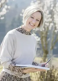 Eine Frau mit kurzen blonden Haaren lächelt, während sie ein Notizbuch und einen Stift hält, draußen vor einem Baum im Hintergrund.