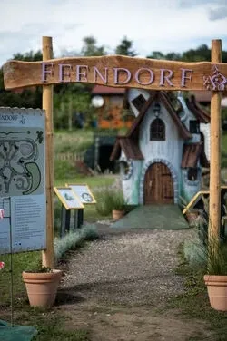Ein Holzschild mit der Aufschrift 'Feendorf' führt zu einem kleinen, verspielten Häuschen mit einer braunen Tür und einem grünen Rasen davor.