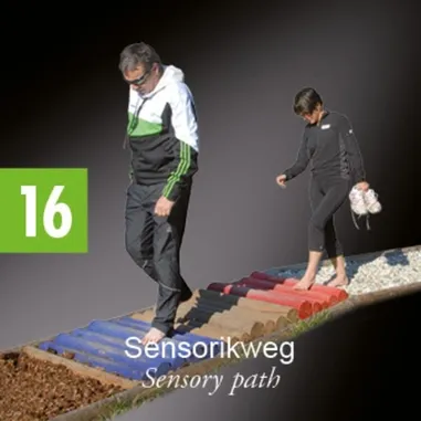 Ein erwachsener Mann und eine Frau gehen auf einem sensorischen Pfad. Der Mann trägt eine Brille und eine weiß-grüne Jacke. Die Frau ist barfuß und trägt ein schwarzes Outfit.