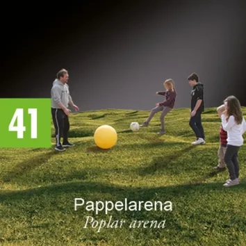 Eine Gruppe von Menschen spielt Fußball auf einem grünen Feld. Ein Mann steht und beobachtet, während ein Mädchen den Ball tritt. Ein Junge steht neben ihr und ein anderes Mädchen schaut auf den Ball.