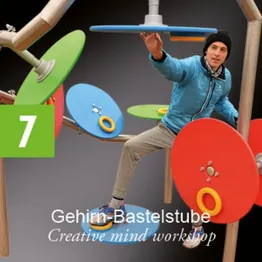 Ein Mann in einer blauen Jacke und grauen Hose steht auf einer Plattform mit einer bunten, interaktiven Kunstinstallation hinter ihm.