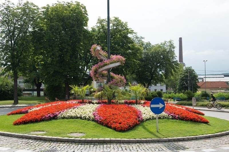 In der Mitte der Straße befindet sich ein Blumenbeet mit roten, weißen und rosa Blumen. Die Blumen sind in Form eines Herzens angeordnet. In der Mitte des Blumenbeets stehen grüne Pflanzen. Eine Person fährt mit dem Fahrrad auf der Straße. Bäume, Straßenlaternen und ein Gebäude befinden sich in der Umgebung.