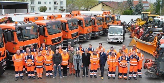 Eine Gruppe von Menschen in orangefarbenen Uniformen steht vor einer Reihe orangefarbener Lastwagen und Maschinenfahrzeuge. Dahinter sind Gebäude und Bäume zu sehen.