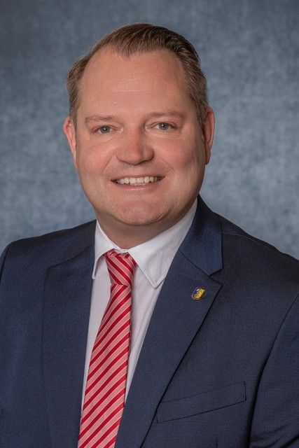 Ein Mann in einem dunkelblauen Anzug und einem rot gestreiften Krawatte lächelt und posiert für ein Foto vor einem grauen Hintergrund.