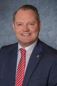Ein Mann in einem dunkelblauen Anzug und einem rot gestreiften Krawatte lächelt und posiert für ein Foto vor einem grauen Hintergrund.