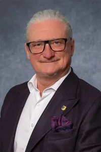 Ein Mann mit weißem Haar und einem Schnurrbart, der eine Brille trägt, ein weißes Hemd und einen dunklen Blazer mit einem lila Einstecktuch und einer gelben Anstecknadel am linken Revers.
