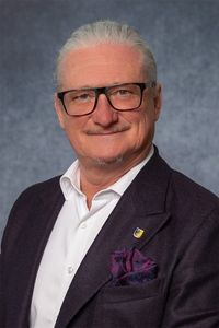 Ein Mann mit weißem Haar und einem Schnurrbart, der eine Brille trägt, ein weißes Hemd und einen dunklen Blazer mit einem lila Einstecktuch und einer gelben Anstecknadel am linken Revers.