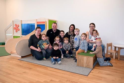 Eine Gruppe von Erwachsenen und Kindern posiert für ein Foto in einem Spielzimmer. Zwei Erwachsene sitzen auf dem Boden mit Kindern um sie herum, während andere dahinter stehen.