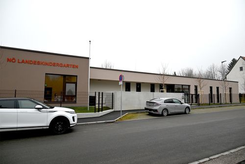 Ein weißer SUV und ein grauer Sedan sind vor einem Gebäude geparkt, das mit 'KINDERGARTEN' beschriftet ist und einen Fahnenmast hat.