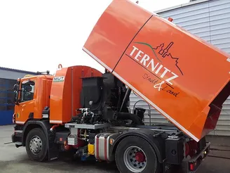 Ein orangefarbener Lastwagen mit dem Wort Terniitz und Stadt und Land steht vor einem Gebäude. Der Lastwagen hat einen großen Container am Heck.