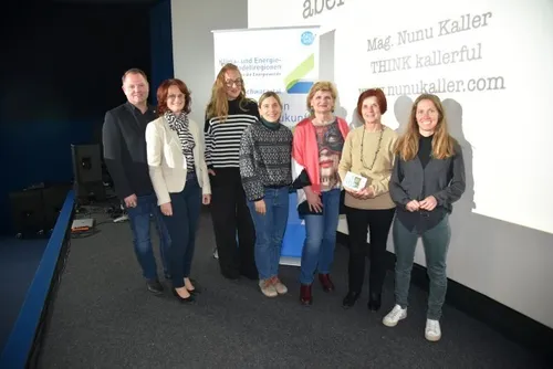 Eine Gruppe von Menschen steht vor einer weißen Wand mit Werbung. Sie lächeln und scheinen für ein Foto zu posieren. Die Wand hat ein Banner mit Text und einem Logo.