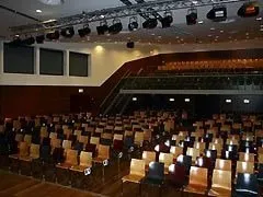 Ein leerer Auditorium mit Reihen von Holzstühlen, die im Theaterstil angeordnet sind. An der Wand befinden sich schwarze Lautsprecher und an der Decke hängende Lampen.