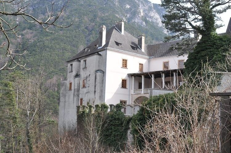 Ein altes weißes Schloss steht auf einem Hügel, umgeben von Bäumen und Pflanzen mit einem bergigen Hintergrund.