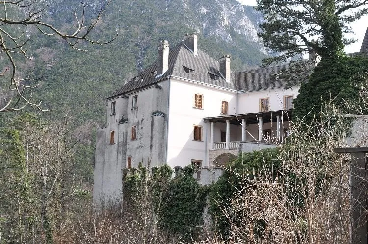 Ein altes weißes Schloss steht auf einem Hügel, umgeben von Bäumen und Pflanzen mit einem bergigen Hintergrund.