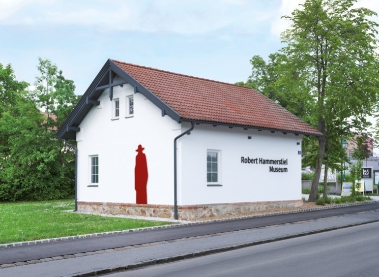 Ein weißes Gebäude mit einer roten Figur an der Wand namens Robert Hammerstiel Museum. Bäume und Gras umgeben das Gebäude.