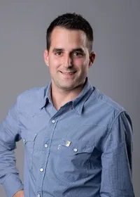 Ein Mann in einem blau karierten Langarmhemd lächelt und posiert für ein Foto mit den Händen auf den Hüften.