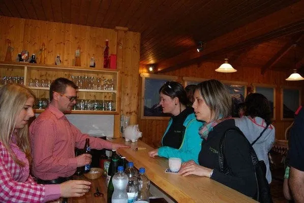 Eine Gruppe von Menschen versammelt sich um eine Holzbar, mit Flaschen, Gläsern und einem Becher auf der Theke. Ein Mann hält eine Flasche und spricht mit einer Frau. Eine andere Frau hält einen Becher, und eine Frau mit einem Schal schaut sie an. Es gibt Regale mit Gläsern, Flaschen und einem schweinförmigen Objekt.